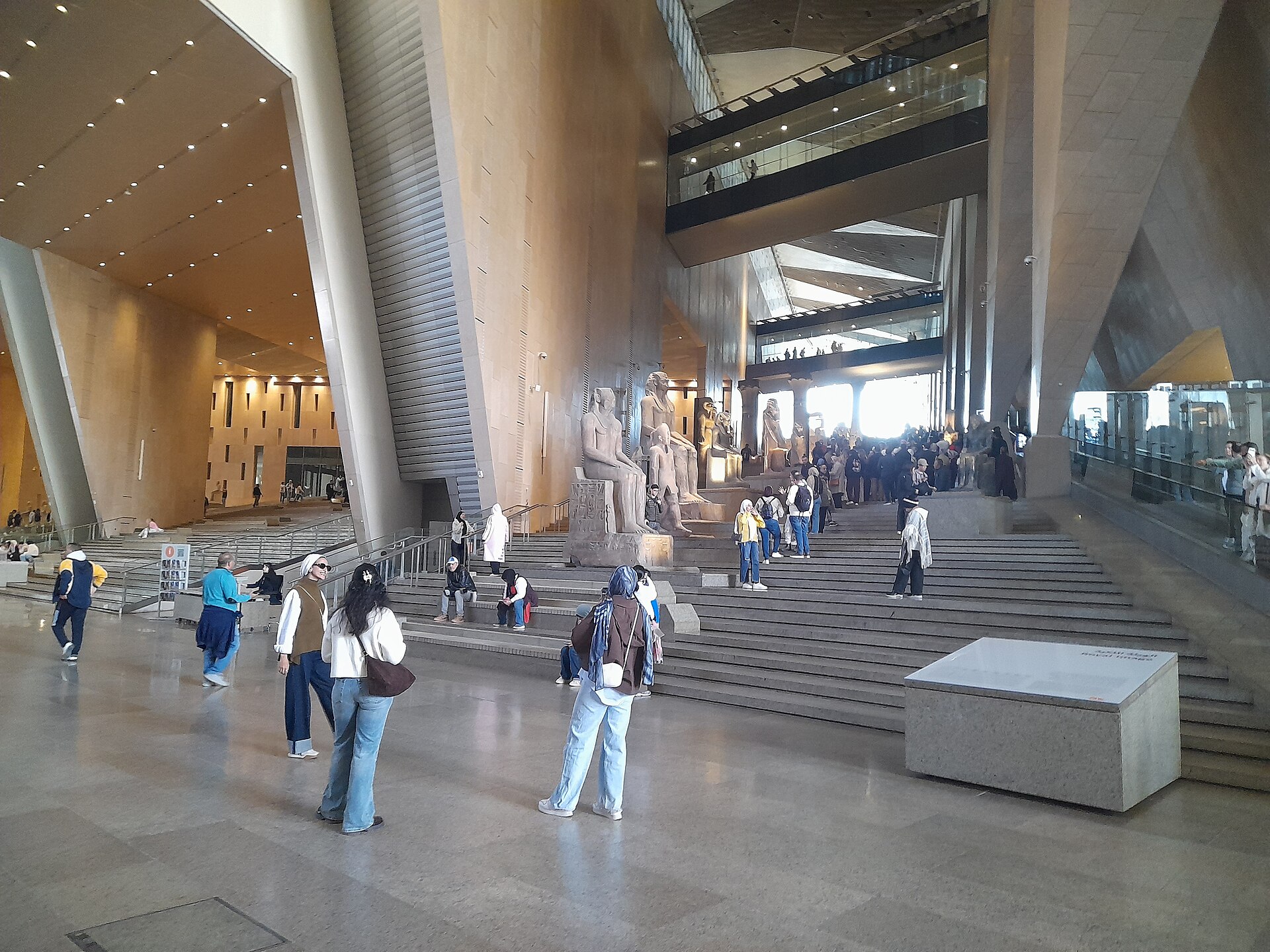 Visitors in the sunlit entrance hall of the Grand Egyptian Museum near Giza, with ancient statues on a wide stone staircase