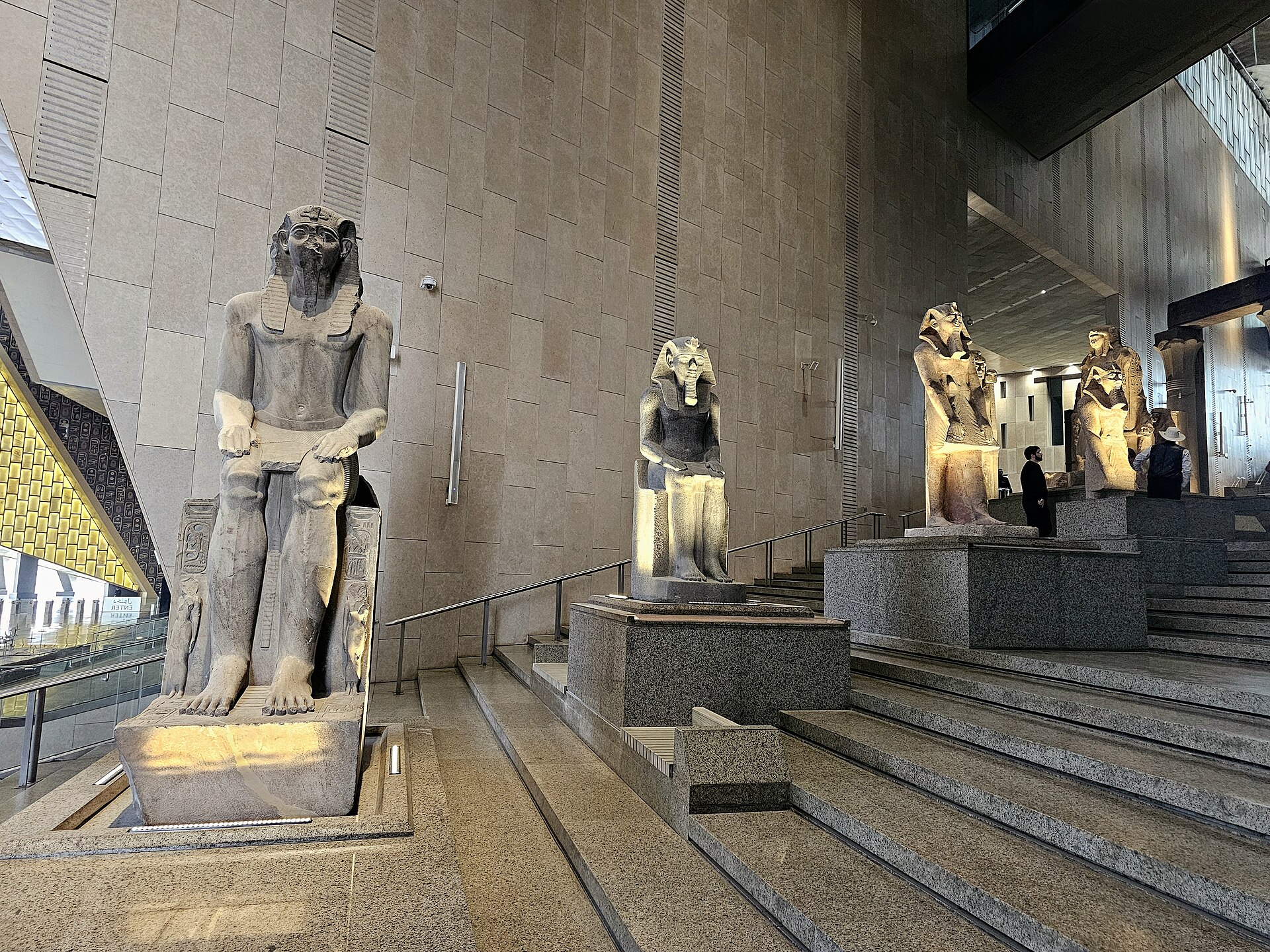 Seated and standing pharaoh statues on stone plinths beside the wide granite Grand Staircase of the Grand Egyptian Museum