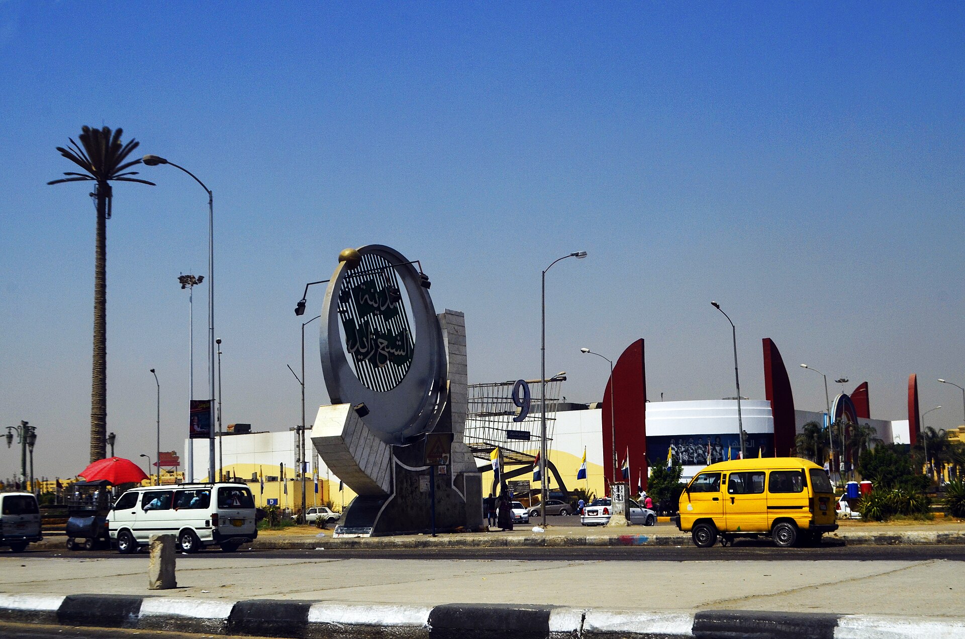 A curved stone monument with Arabic lettering at a roundabout entrance to Sheikh Zayed City, with a yellow minibus, palm tree and a shopping centre behind