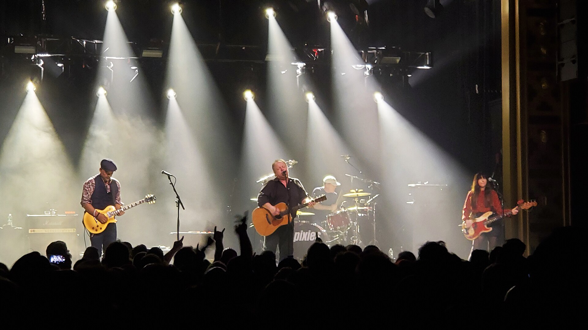 The Pixies on a dark stage under beams of white light, with a guitarist, a singer with an acoustic guitar, a drummer and a bassist, and the silhouettes of the audience in the foreground