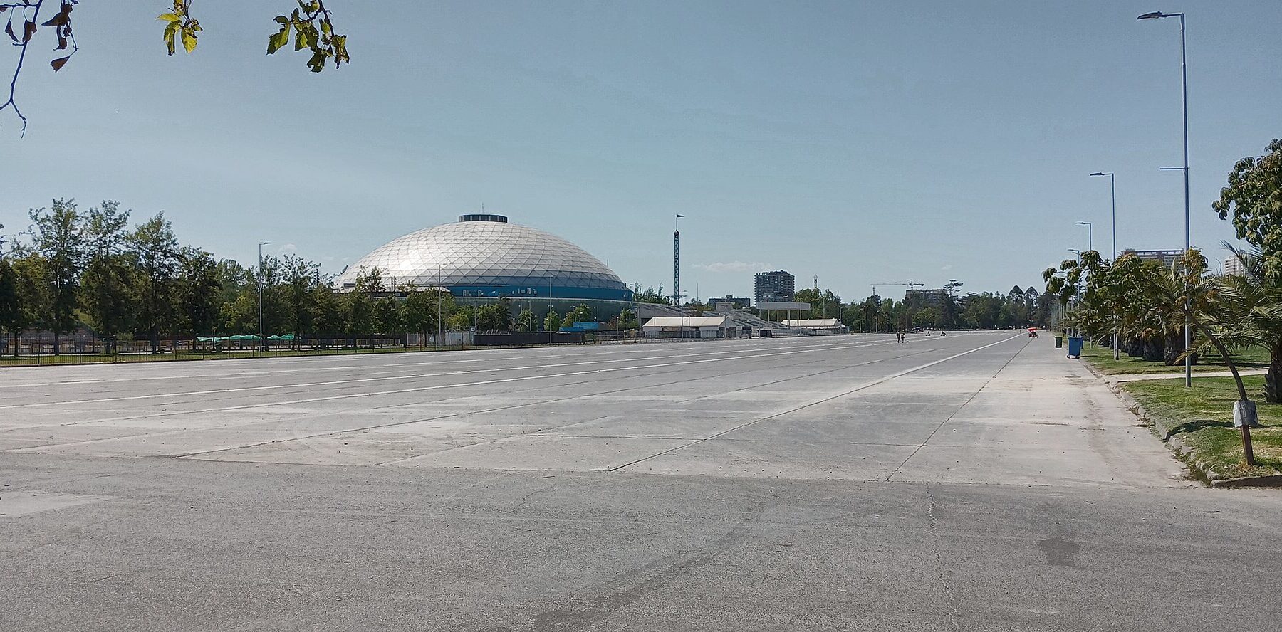 A wide paved esplanade in Parque O'Higgins in Santiago with the white dome of the arena, trees and a lamp-lined avenue under a blue sky