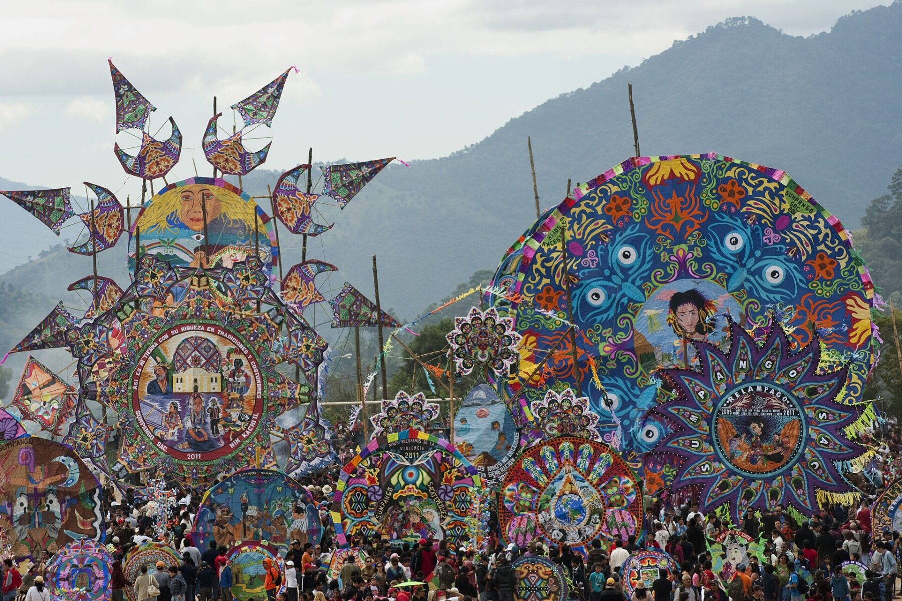 Dozens of huge round tissue-paper kites in bright colours stand on poles above a dense crowd, with forested mountains behind
