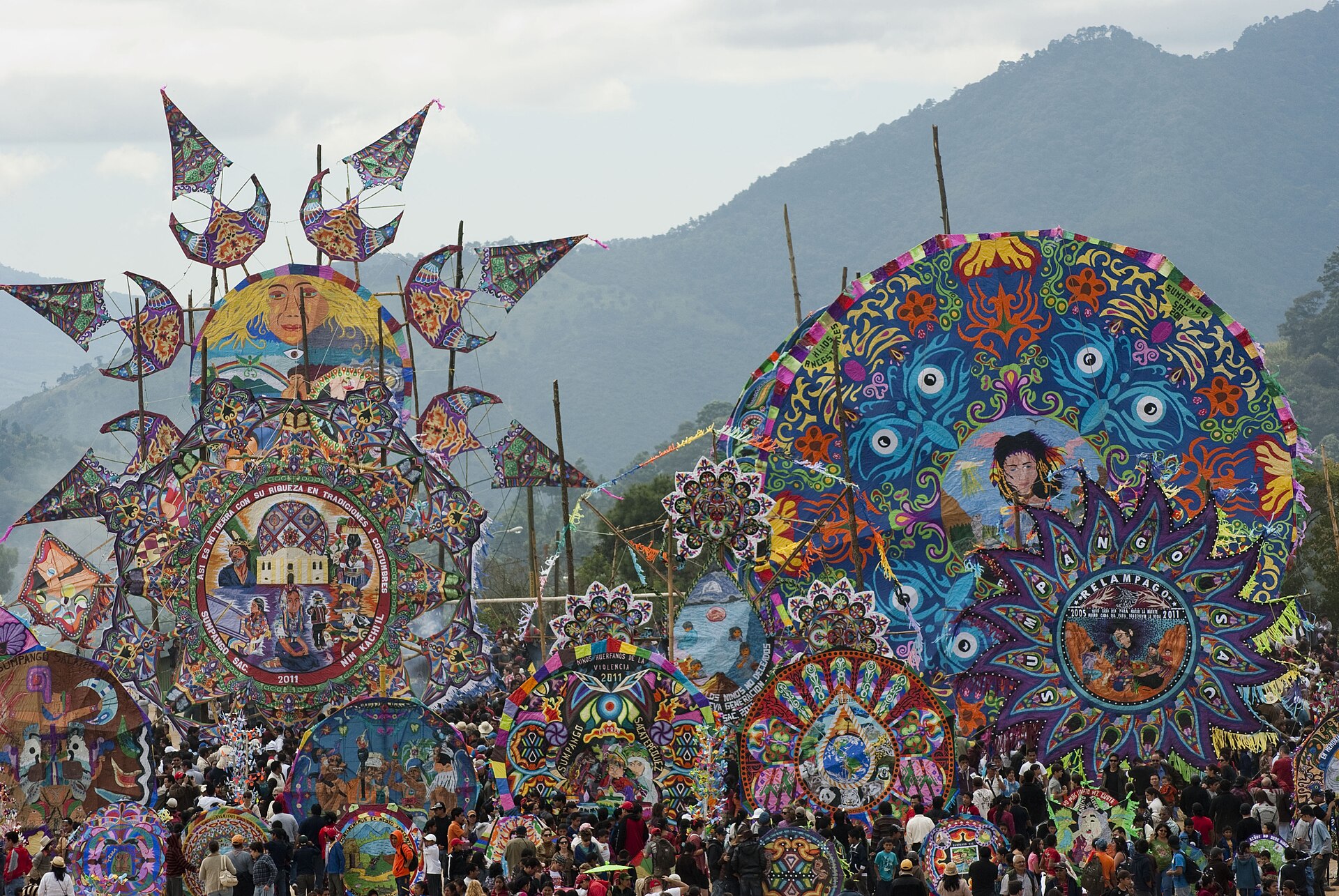 Dozens of huge round tissue-paper kites in bright colours stand on poles above a dense crowd, with forested mountains behind