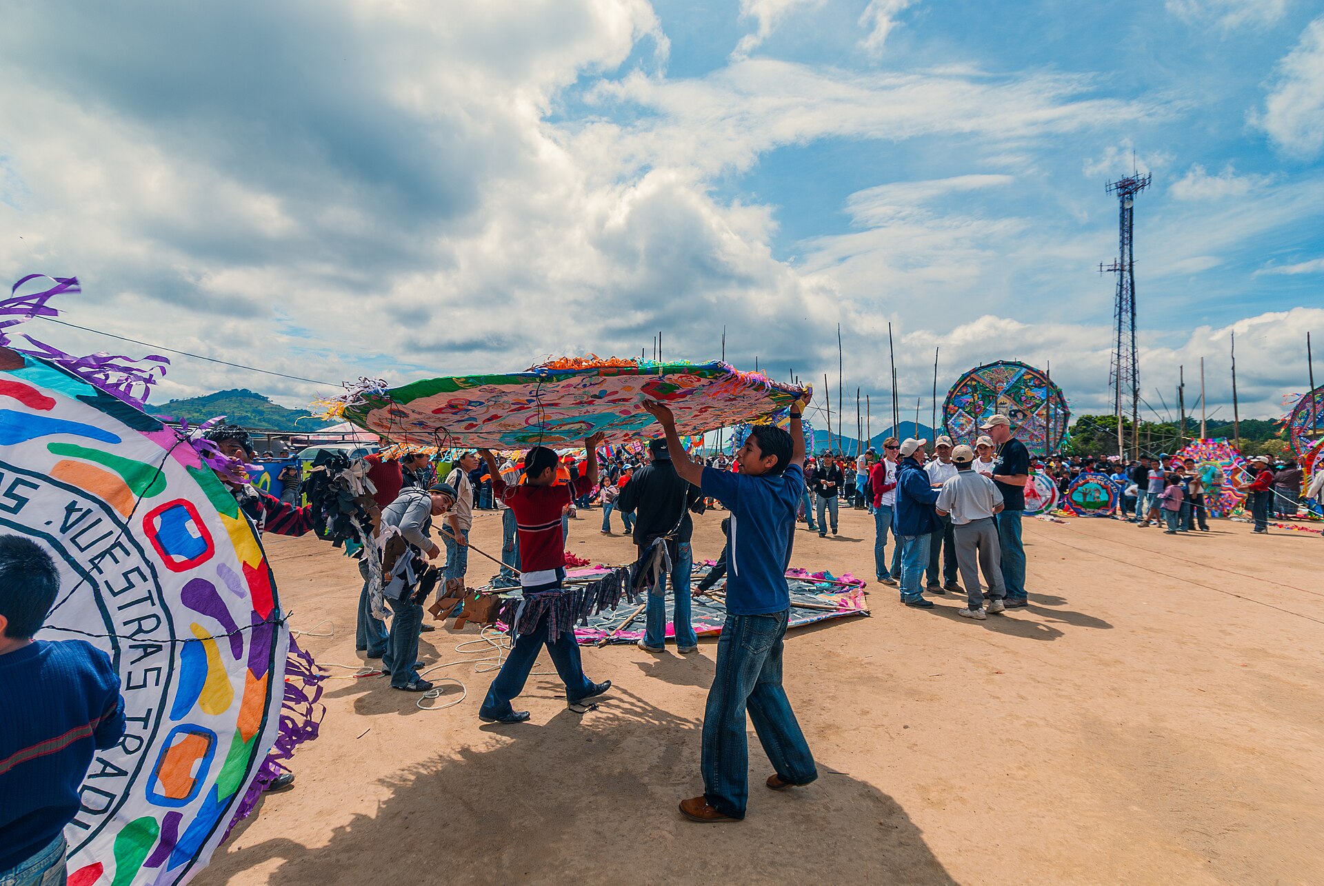 Young men carry a large round kite over their heads across a sandy field, with other giant kites on poles and a telecom mast in the distance under a cloudy sky