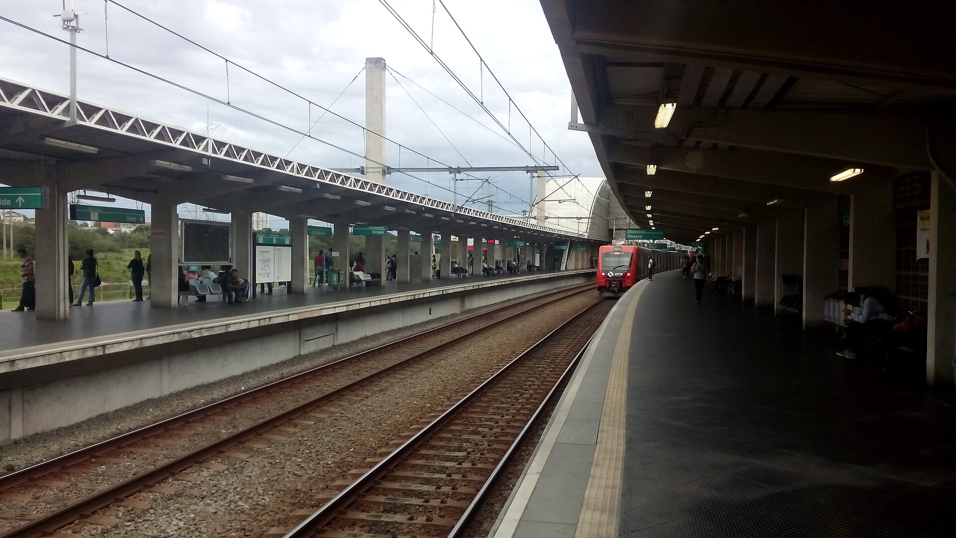 Covered platforms and tracks at Jurubatuba commuter rail station in São Paulo, with a red train standing at the far end under a cloudy sky