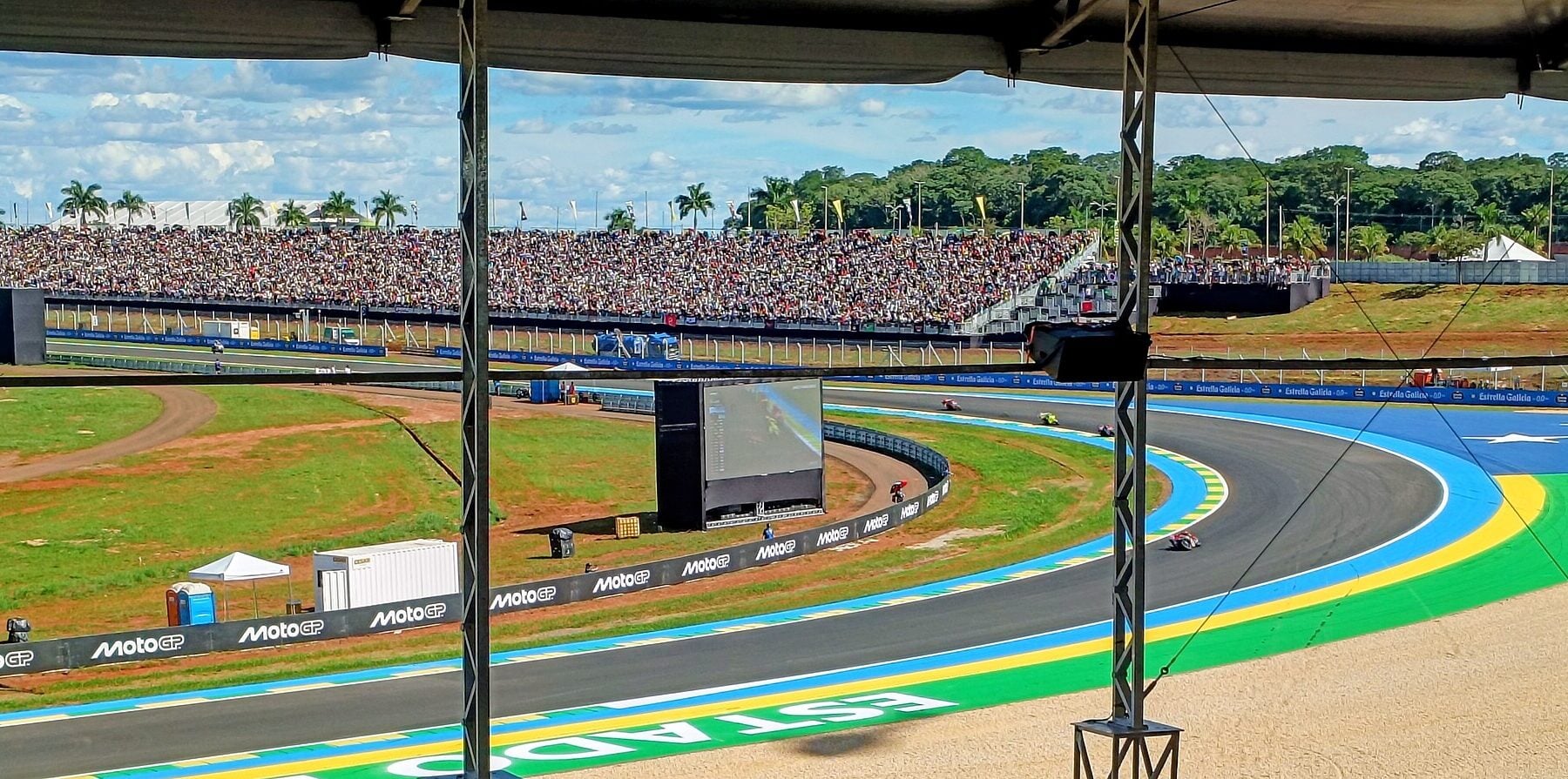 MotoGP riders on a blue-and-green kerbed corner of the Goiânia circuit, seen from under a covered stand, with a packed grandstand, a big screen and palm trees behind