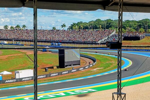 MotoGP riders on a blue-and-green kerbed corner of the Goiânia circuit, seen from under a covered stand, with a packed grandstand, a big screen and palm trees behind