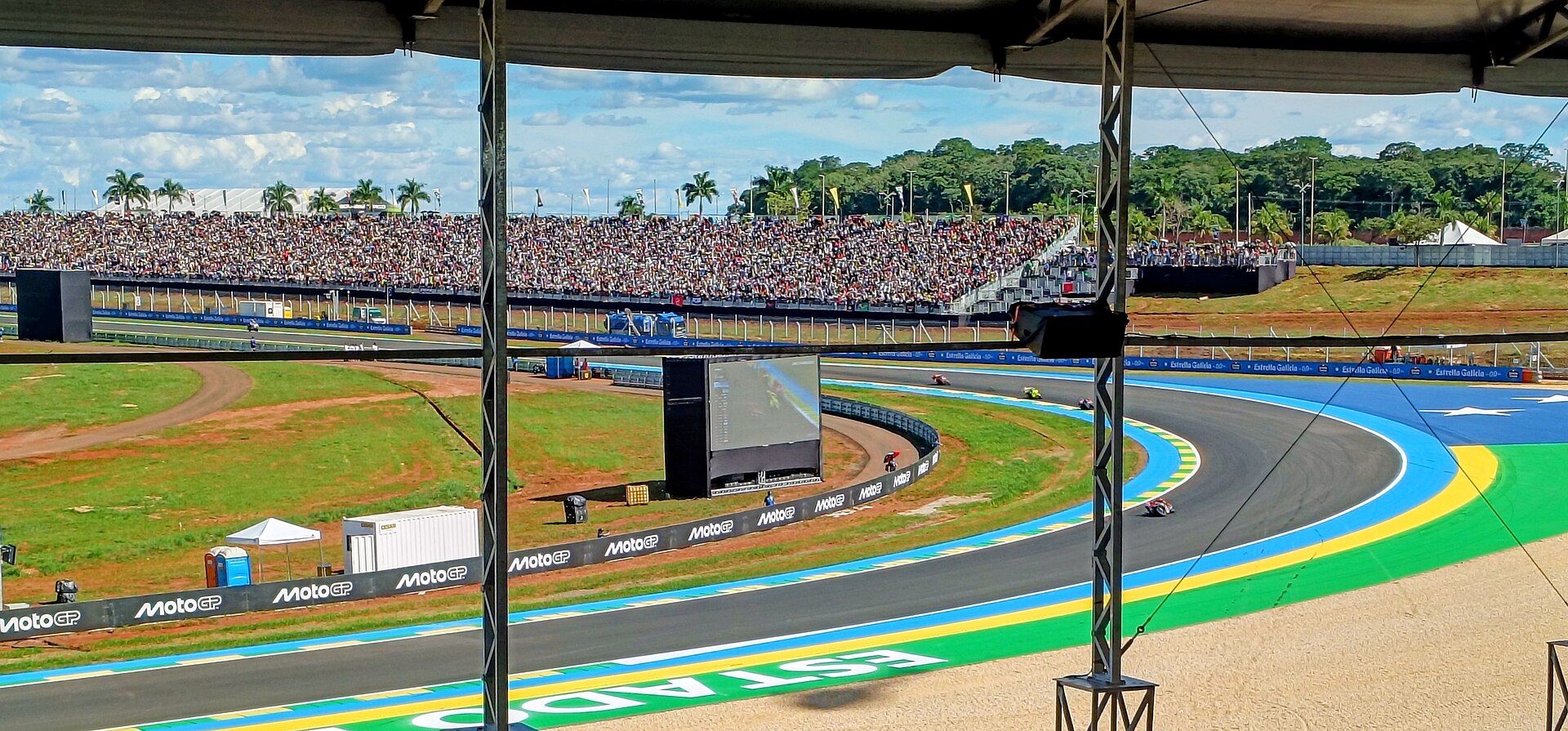 MotoGP riders on a blue-and-green kerbed corner of the Goiânia circuit, seen from under a covered stand, with a packed grandstand, a big screen and palm trees behind