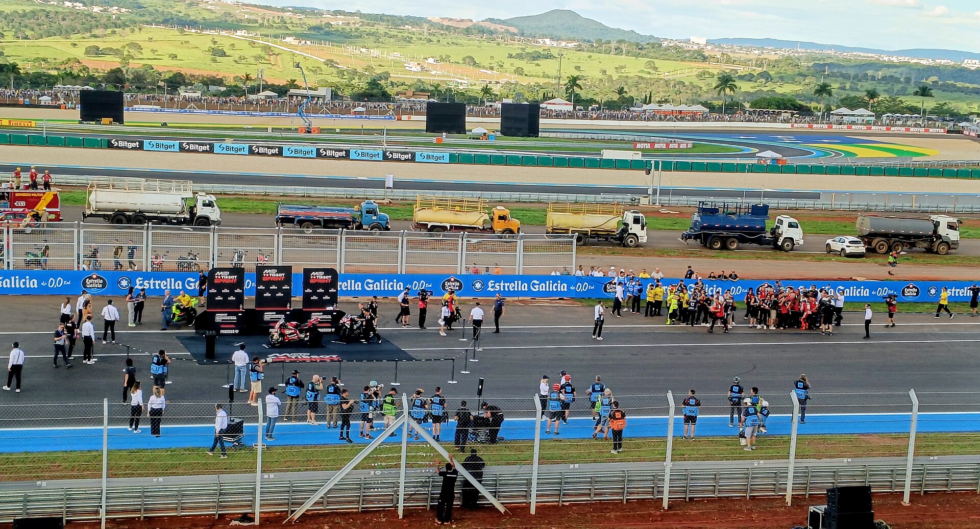 Motorcycles parked beside MotoGP Tissot Sprint boards on the main straight of the Goiânia circuit, with team crews, photographers, Estrella Galicia banners, water trucks and green hills behind