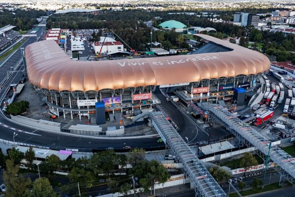 Drone view of Estadio GNP Seguros in Mexico City, a stadium with a curved copper-coloured roof reading Vivir es increíble, ringed by the Autódromo Hermanos Rodríguez racetrack and footbridges