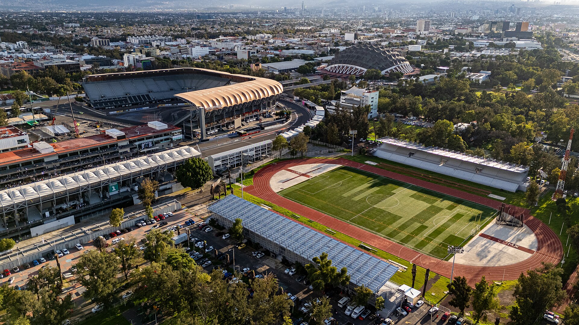 Aerial view of a Mexico City sports complex with a copper-roofed stadium at left, a domed arena behind and an athletics track with a football pitch in the foreground