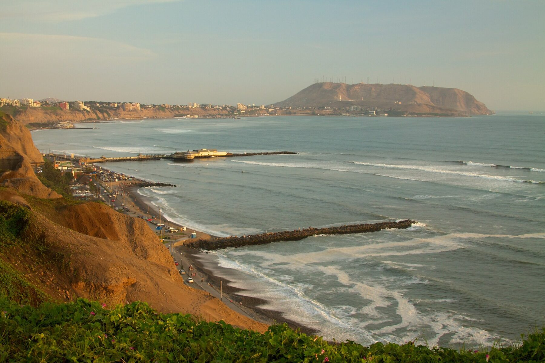 Evening sun on brown coastal cliffs above a curving beach road, with a pier, breakwaters and a large headland across a wide bay