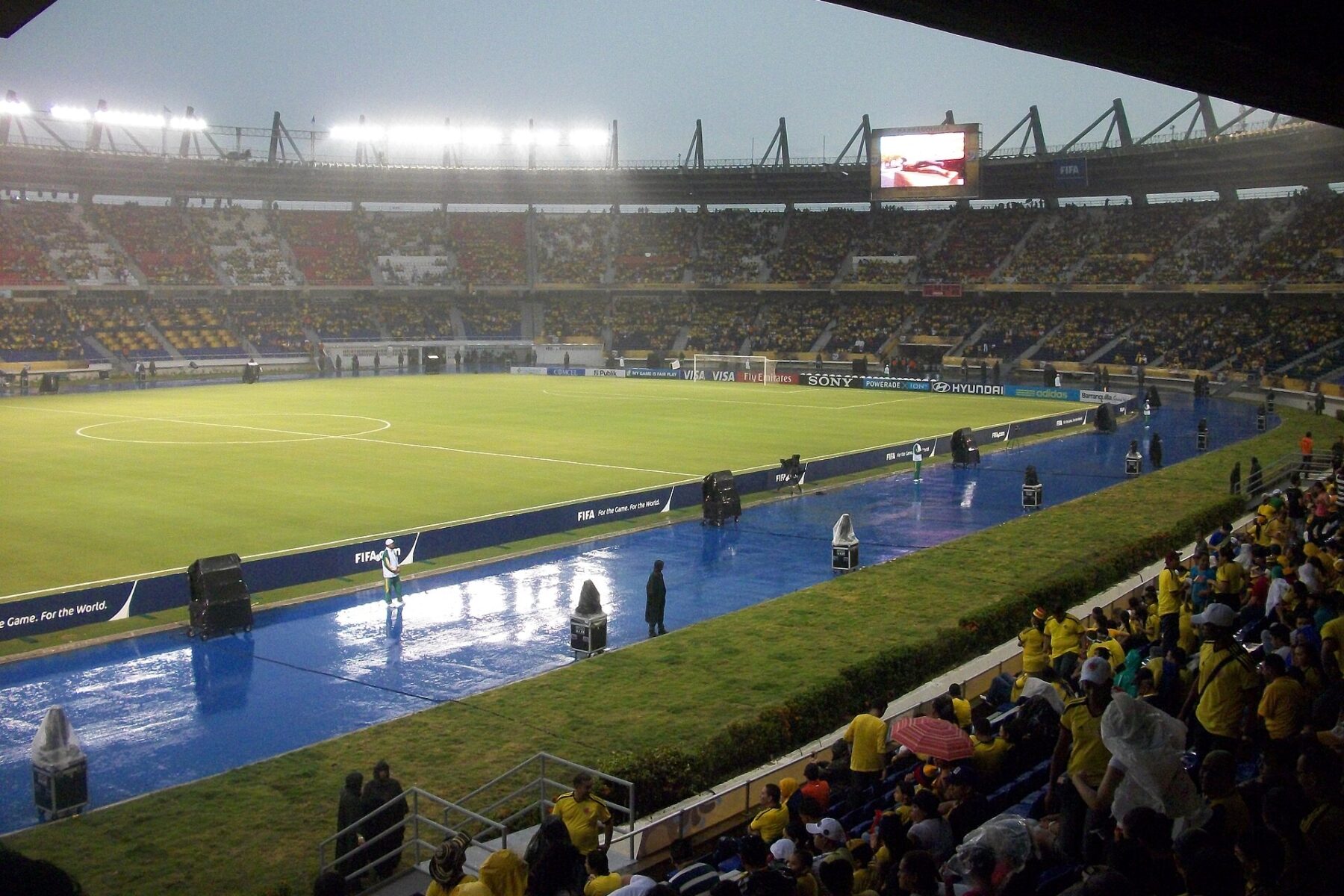 Fans in yellow shirts watch from the stands of the Estadio Metropolitano in Barranquilla as rain falls on the floodlit pitch