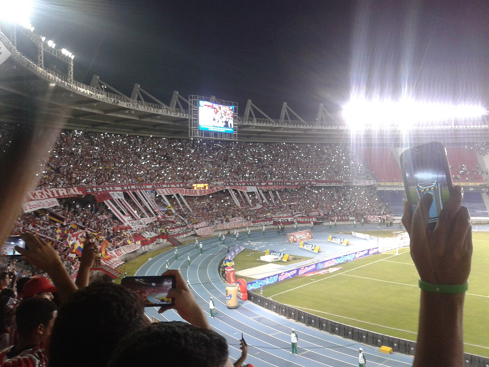 Fans in red and white hold up phone lights in the packed stands of a floodlit stadium at night, with banners along the lower tier