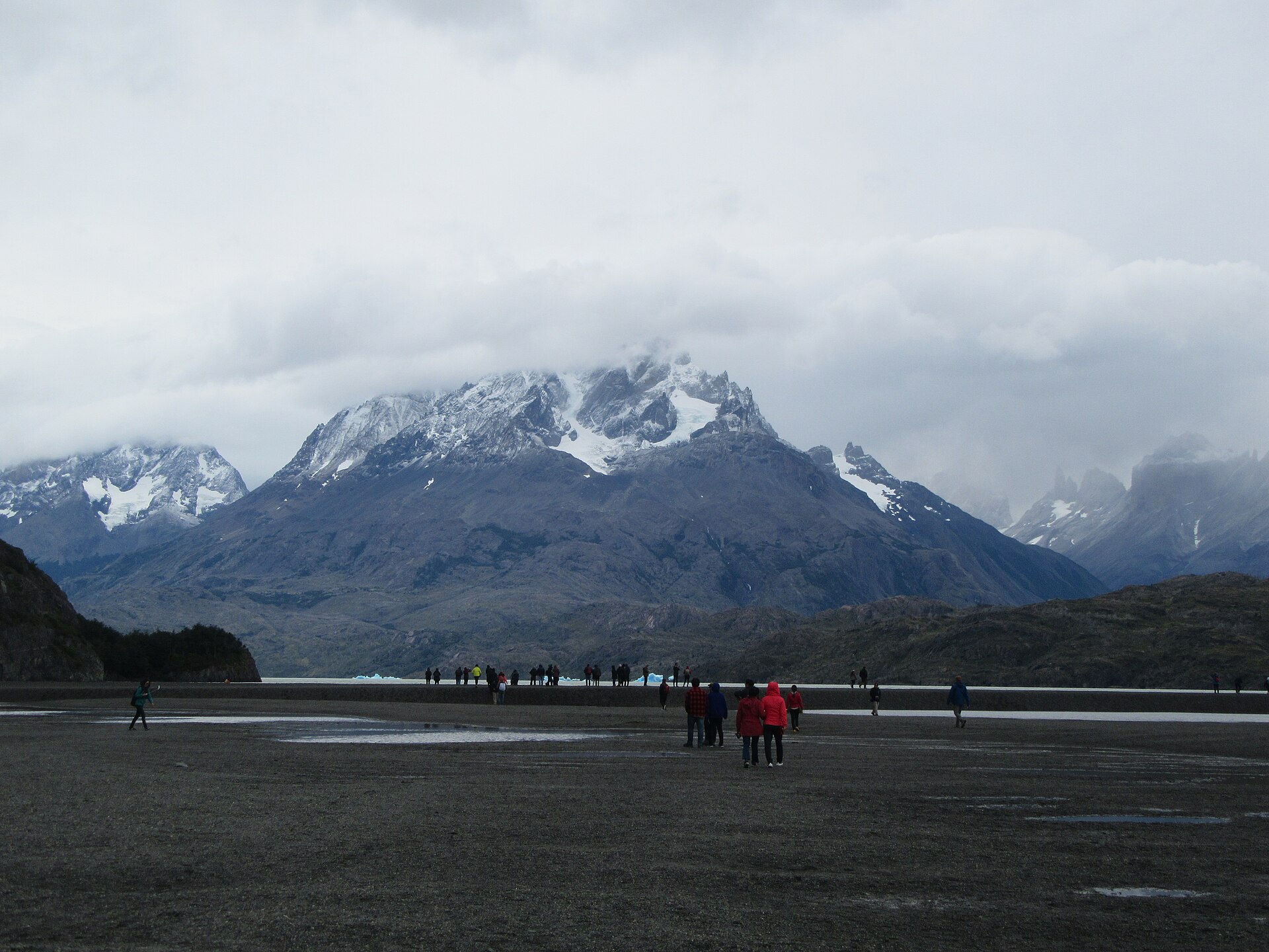 Small groups of visitors in rain jackets walk across a flat grey lakeshore towards floating ice, with snow-capped mountains half hidden in cloud behind