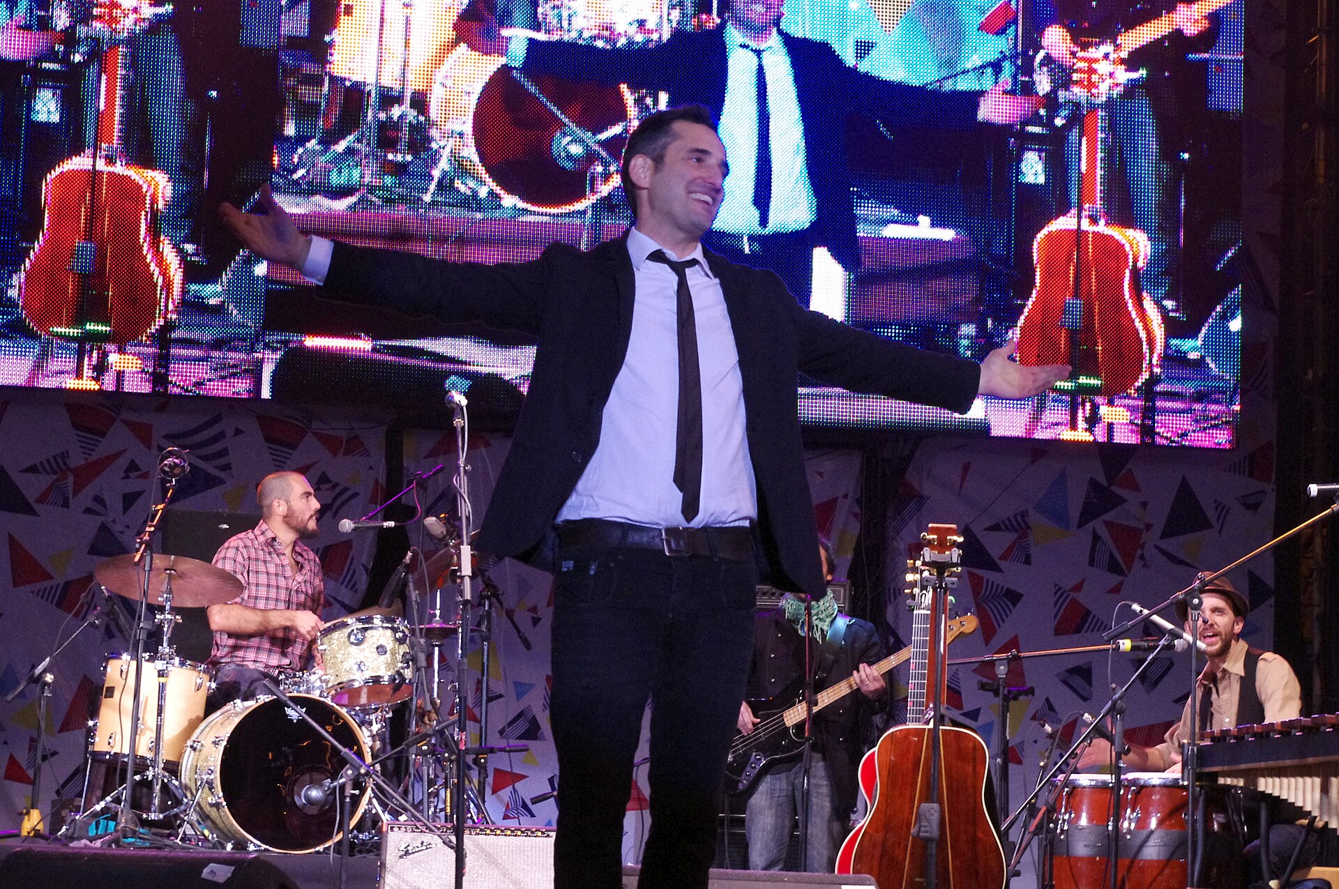 A smiling man in a dark suit and thin tie stands on stage with his arms spread wide, with a drummer, a bass player, acoustic guitars on stands and a percussionist at congas and vibraphone behind him and a large video screen above