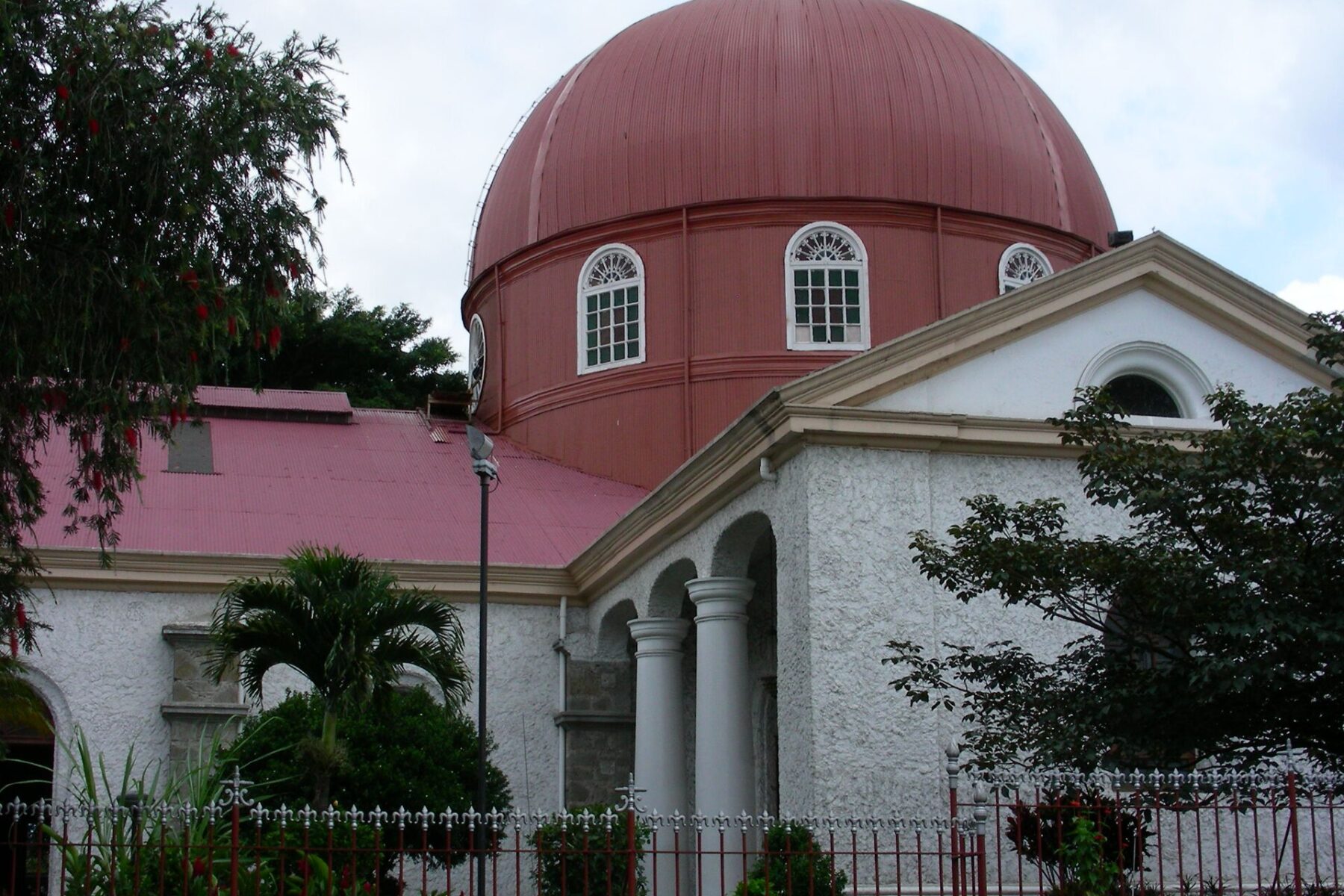 A large red metal dome with arched windows rises above a white church with columns, a red roof, palm trees and a red iron fence