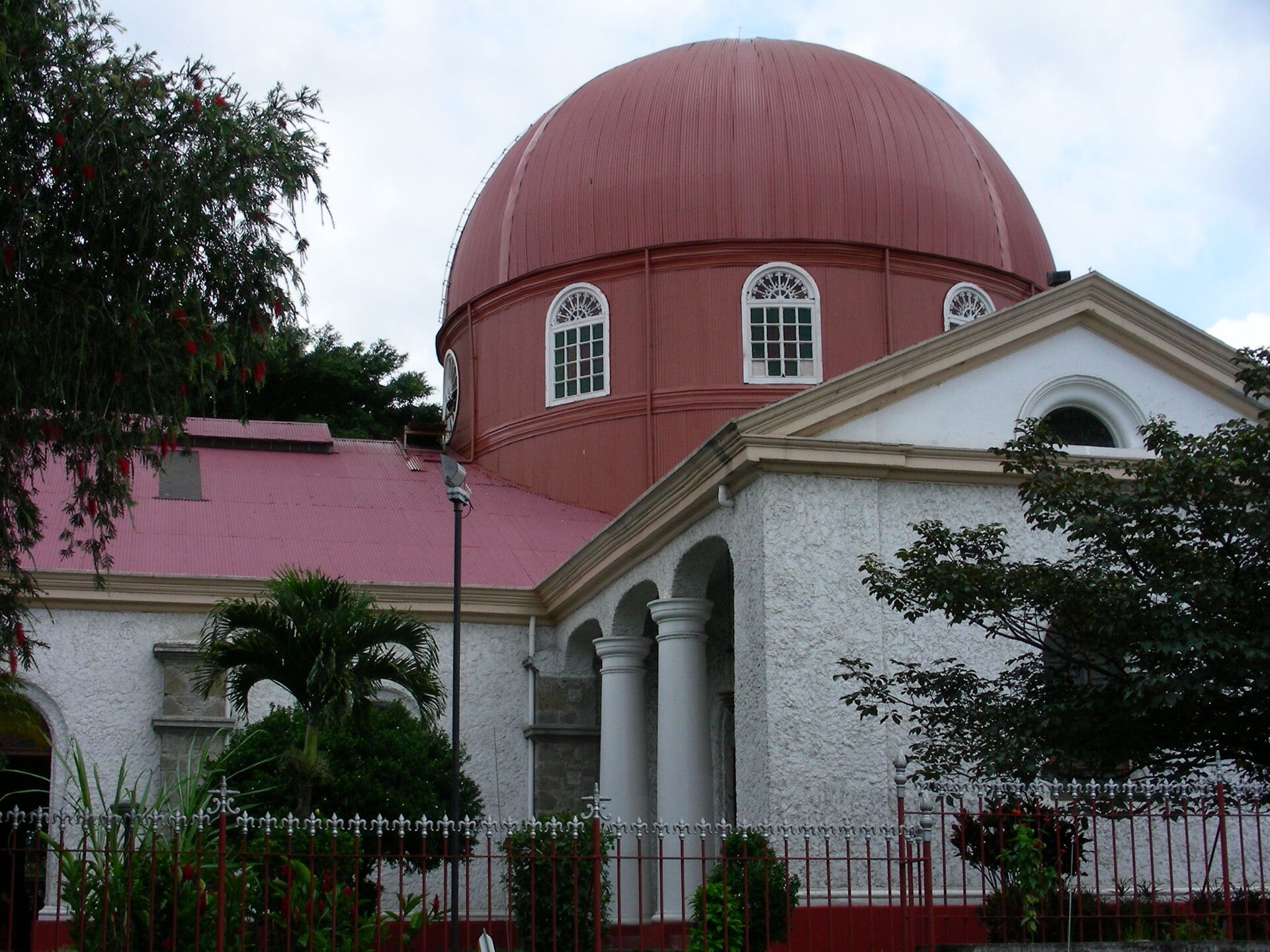 A large red metal dome with arched windows rises above a white church with columns, a red roof, palm trees and a red iron fence