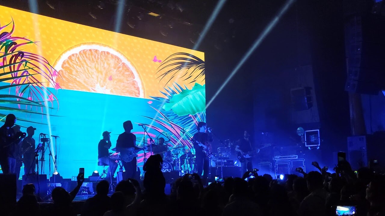 Rawayana performing on a dark stage lit by blue light, in front of a large screen showing an orange slice and palm leaves, with fans filming on phones