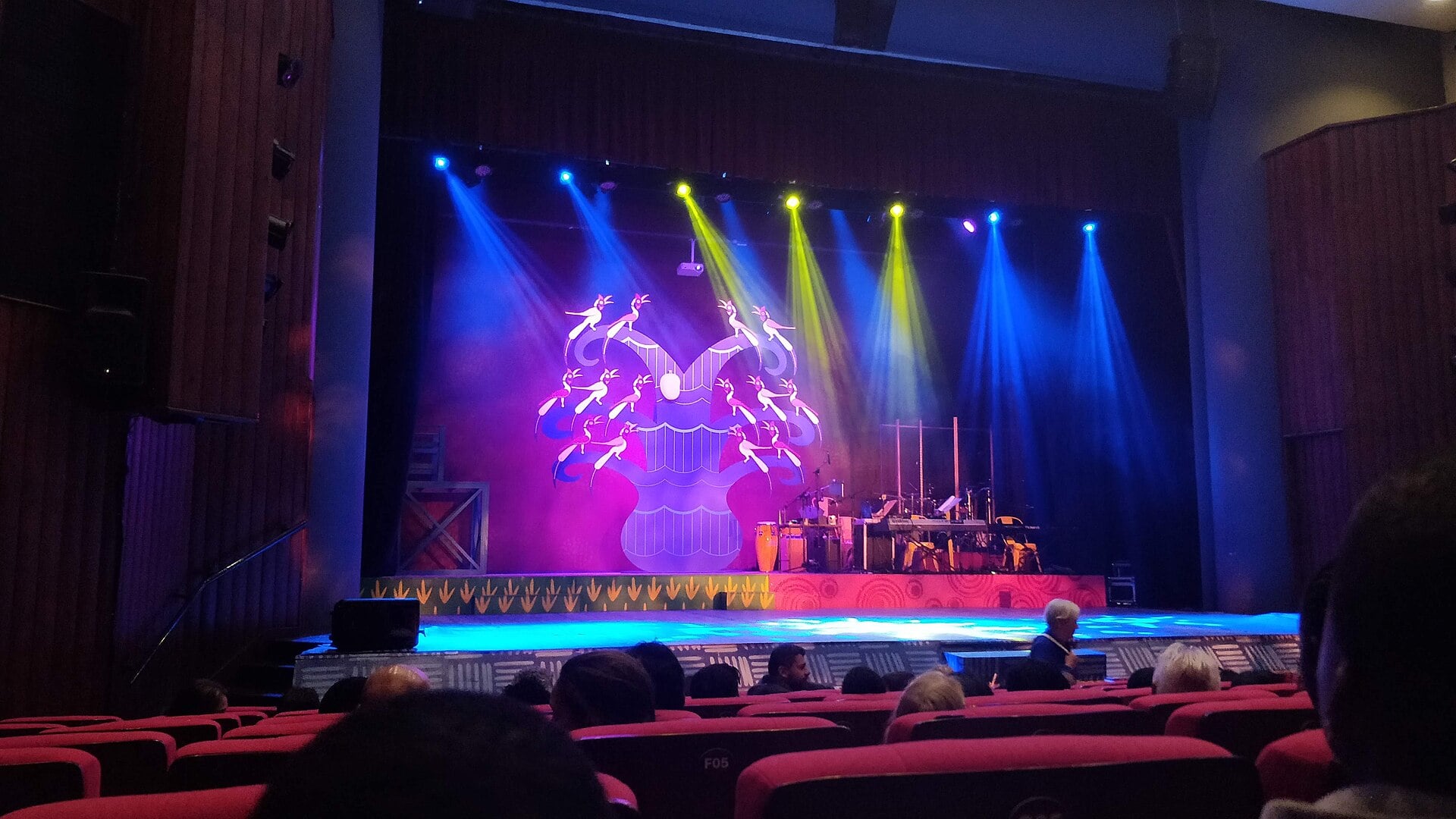 A theatre stage lit by blue and yellow spotlights in front of a stylised tree backdrop with birds, seen over rows of red seats with audience members