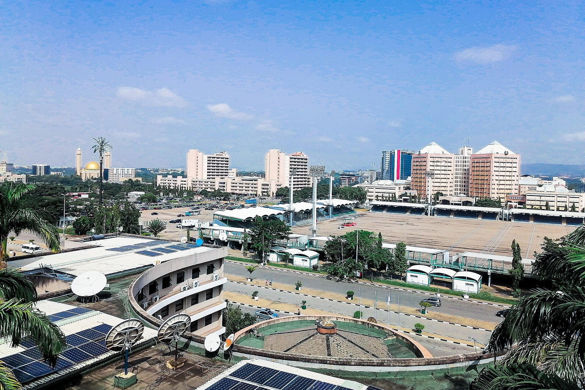 Eagle Square in Abuja seen from a rooftop, with its covered stands and open parade ground, office towers behind and the gold dome of the National Mosque on the left, under a blue sky