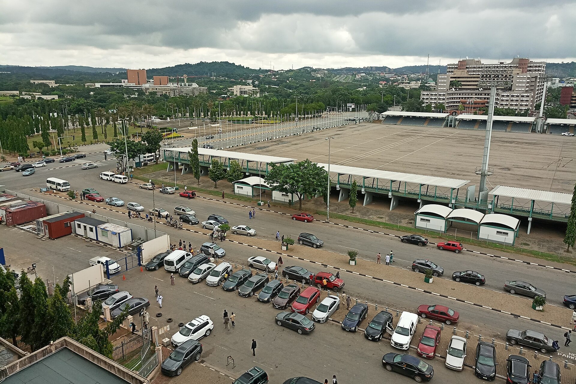 Elevated view of Eagle Square in Abuja, with white-roofed stands along a wide parade ground, a road and a full car park in front and green hills behind under a cloudy sky