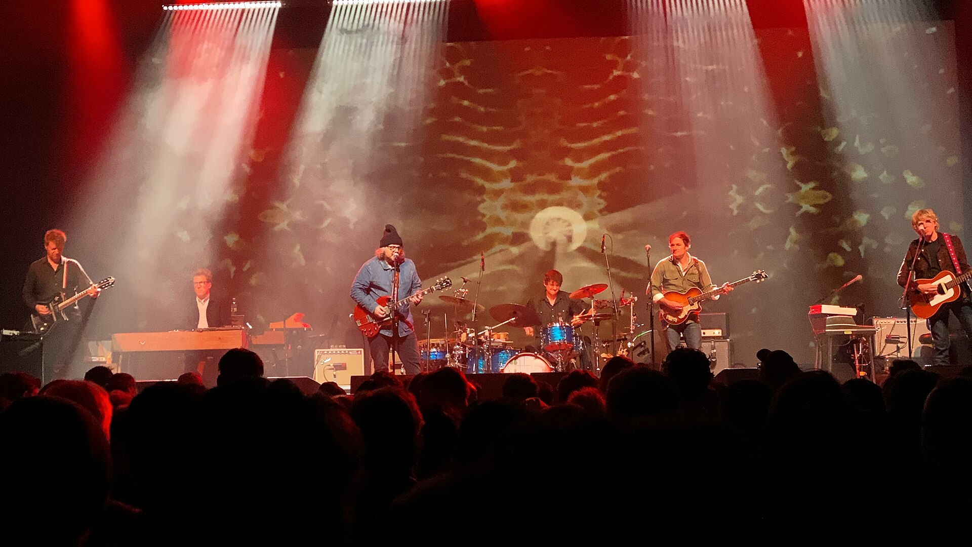 The six members of Wilco performing on a stage lit by red and white spotlights, seen over the heads of the audience