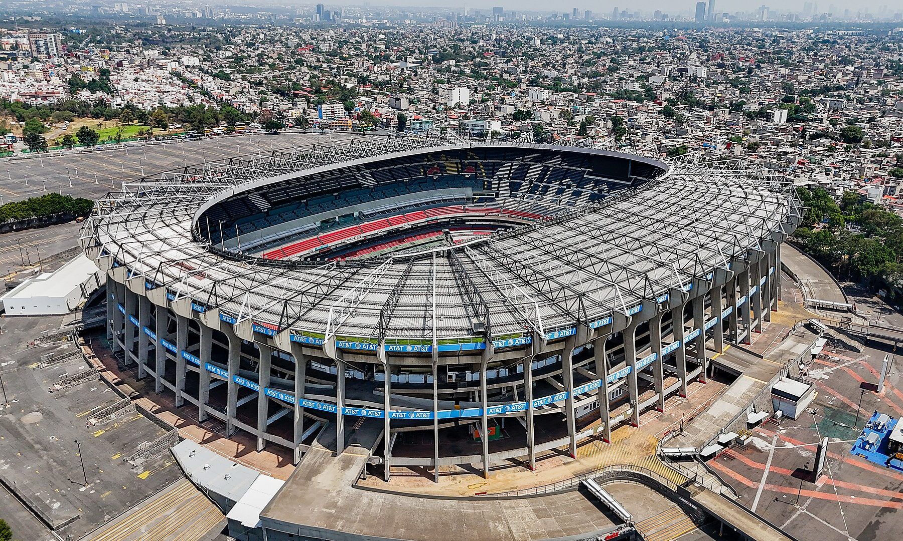 Aerial view of a large oval football stadium with a steel roof ring and blue and red seats, surrounded by a parking area and dense city blocks