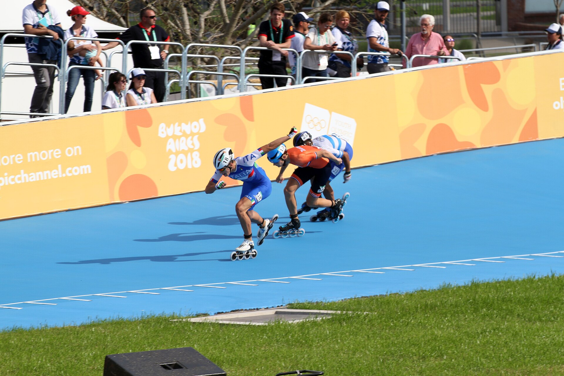 Three inline speed skaters lean into a bend on a blue track in front of spectators behind a Buenos Aires 2018 banner