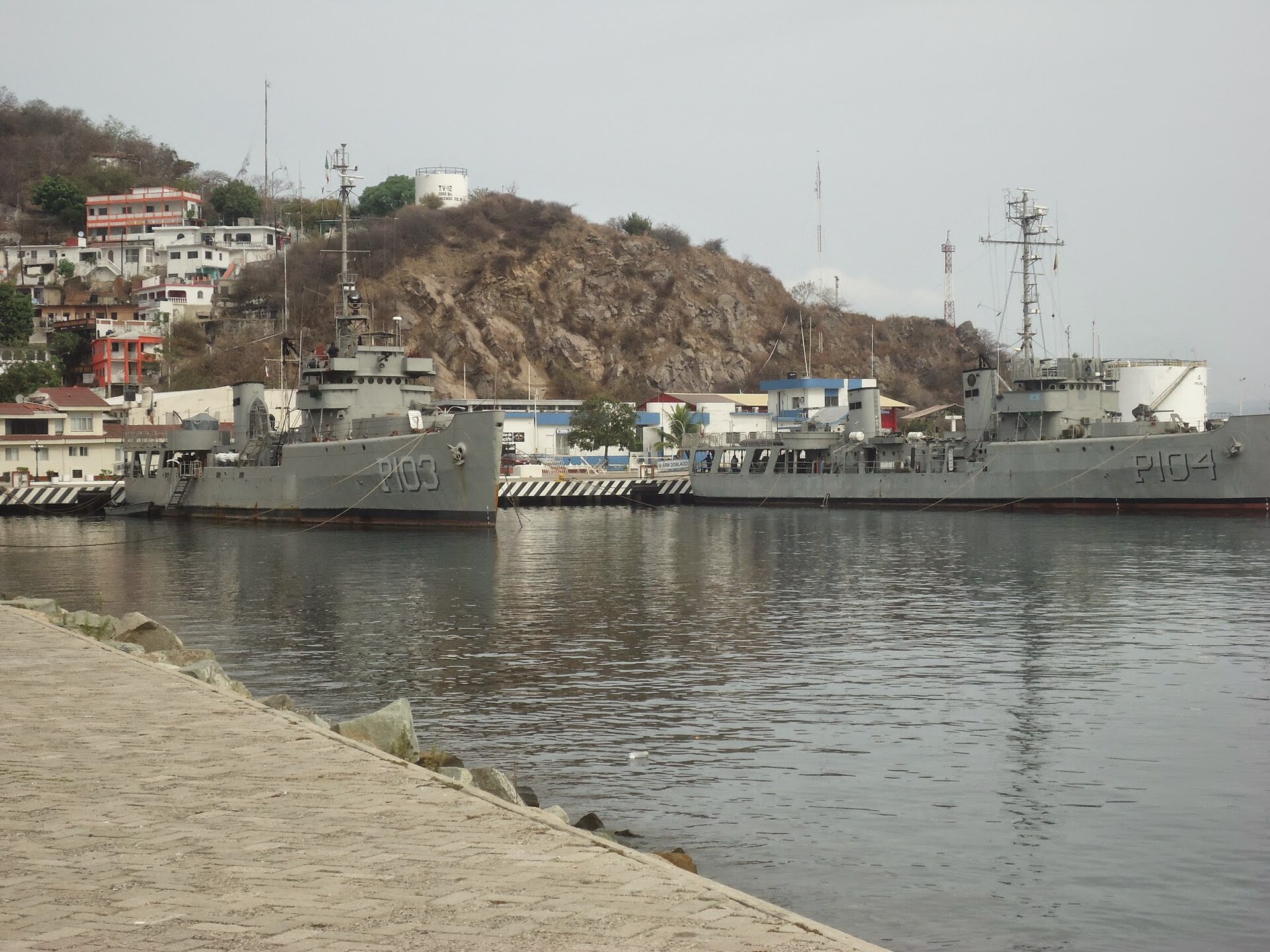 Two grey Mexican Navy patrol ships, hull numbers P103 and P104, moored at a pier below a rocky hill with houses, seen from a stone jetty