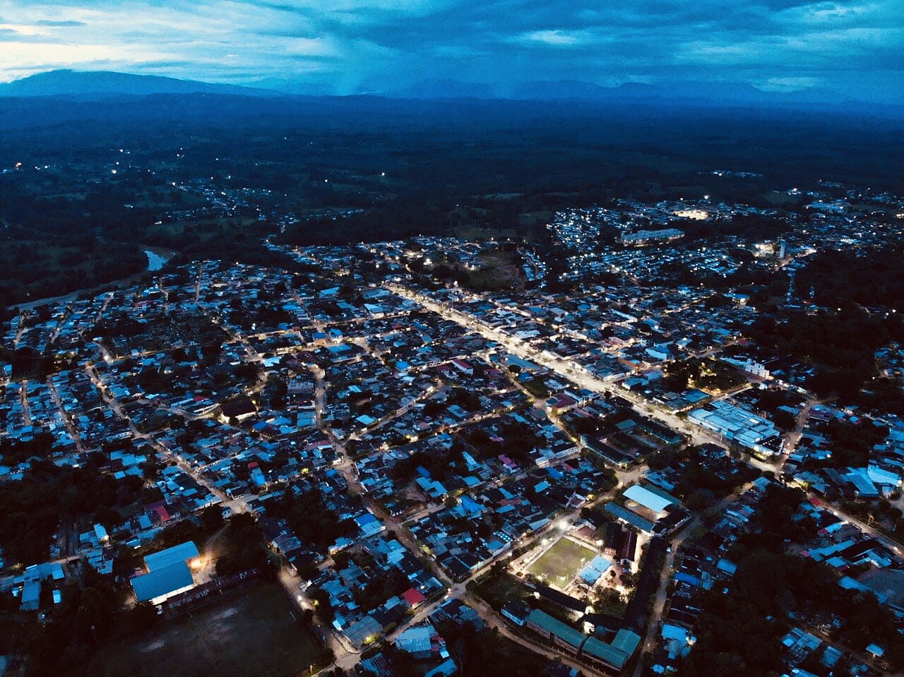 Aerial view of the lit street grid of Tibú at dusk with dark hills and clouds behind