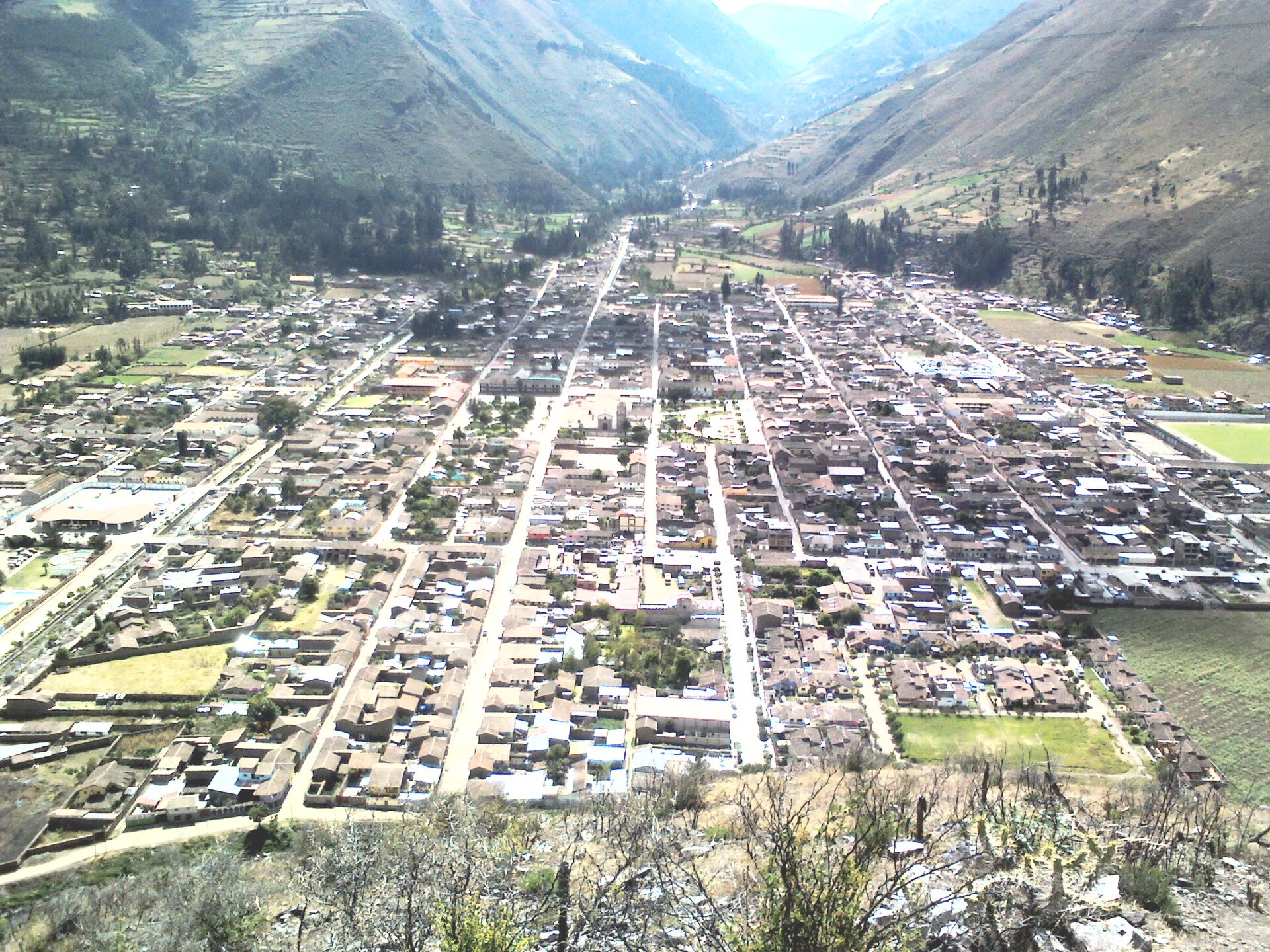 Elevated hillside view of a grid-patterned Andean town with tiled roofs and green fields, set in a narrow valley between steep brown mountains