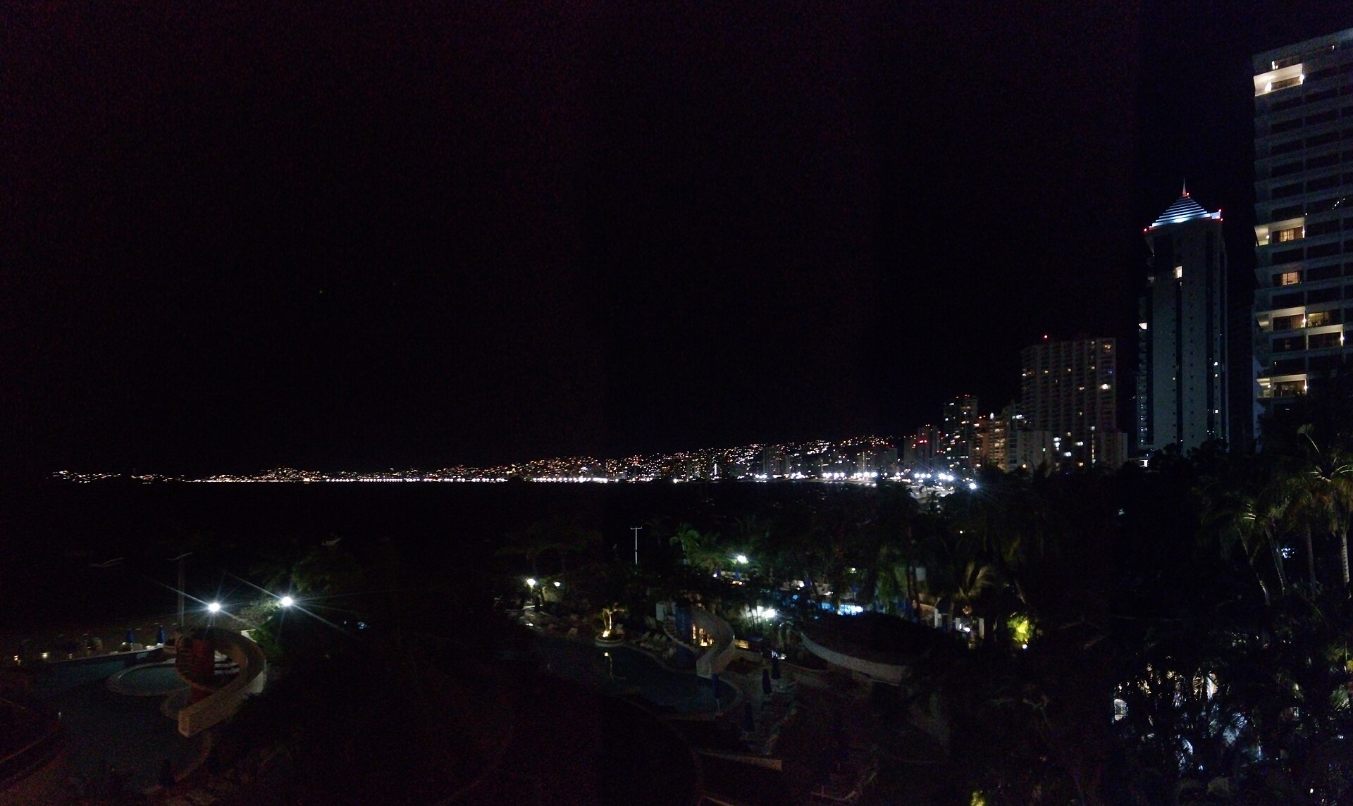 Night view over a dark bay with a line of city lights along the shore, high-rise towers on the right and lit hotel pools in the foreground