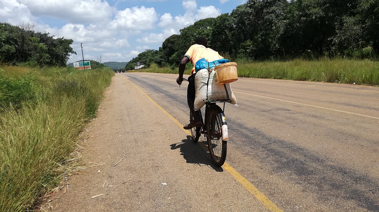 A man cycles along a road with a sack of maize and a bucket on the back of his bicycle