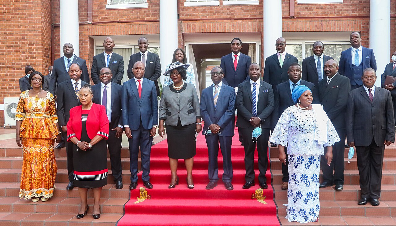 Ministers and officials in formal dress posing for a group photo on red-carpeted steps outside a brick government building in Zambia