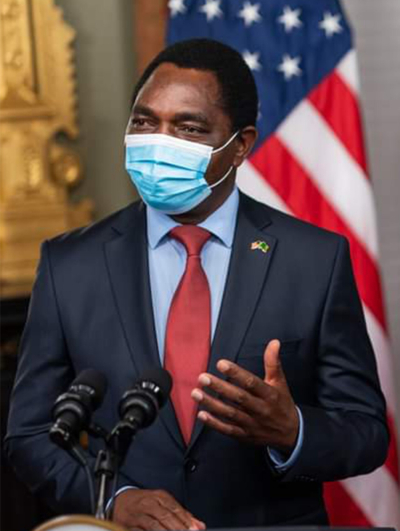Close-up of a man in a dark suit and red tie wearing a blue face mask, speaking at two microphones in front of a US flag