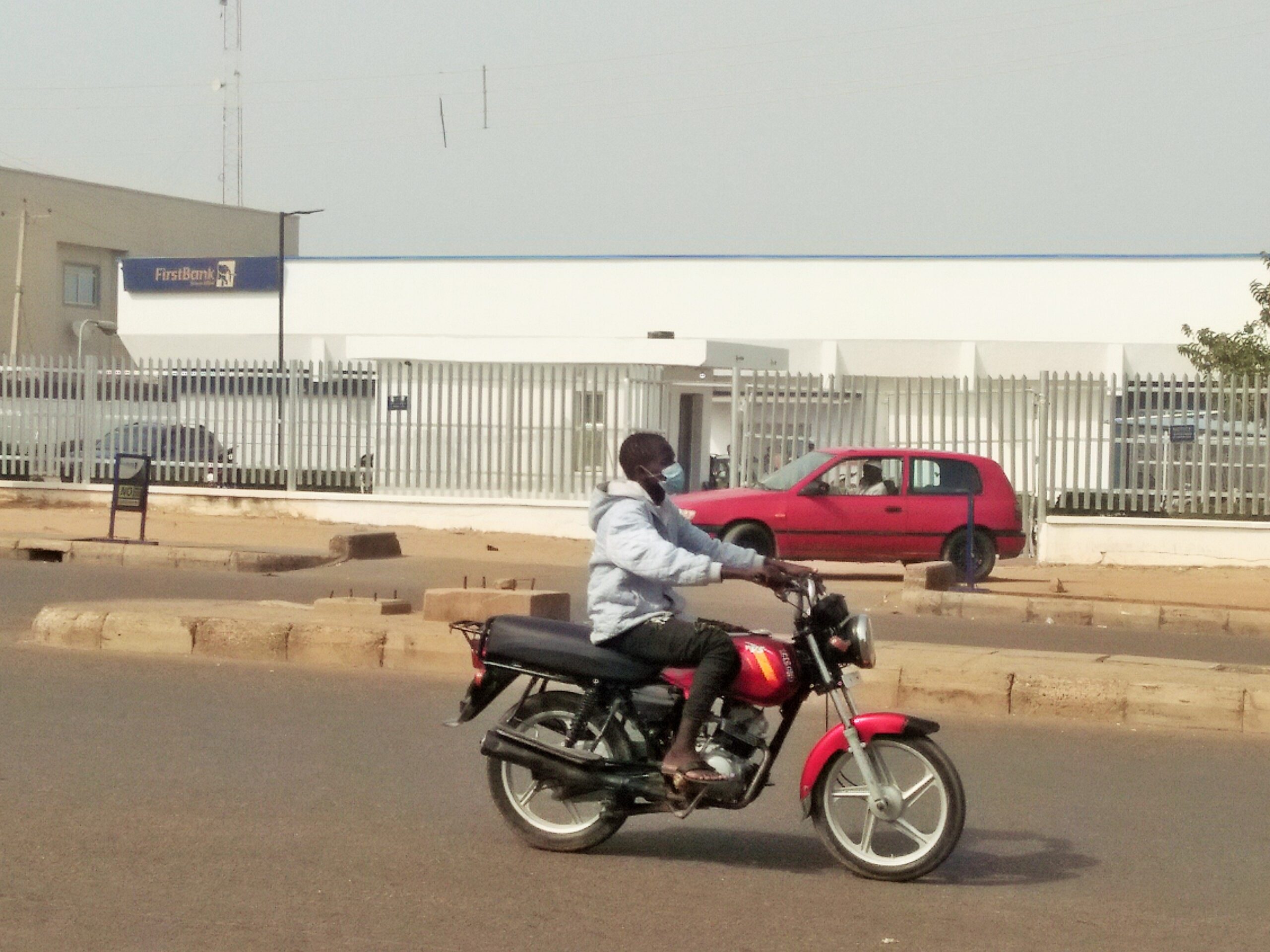 A man on a red motorcycle passes a white bank branch with a blue sign behind a fence on a street in northern Nigeria