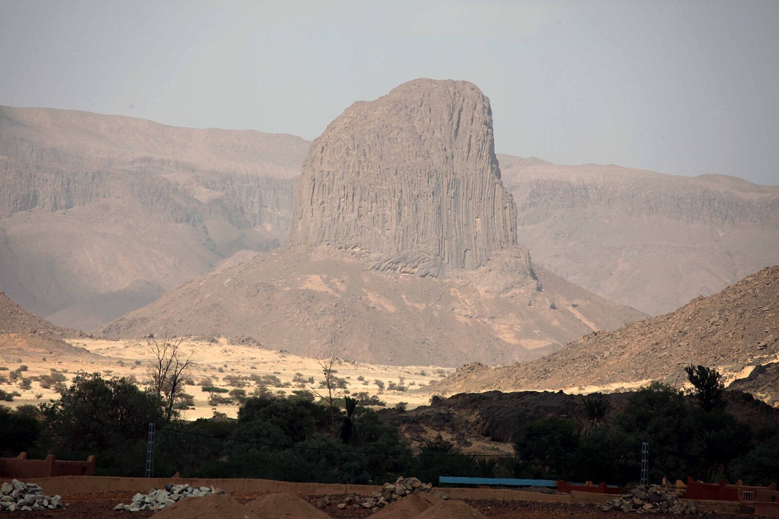 A tall rock tower rising above desert hills and scattered trees in the Sahara