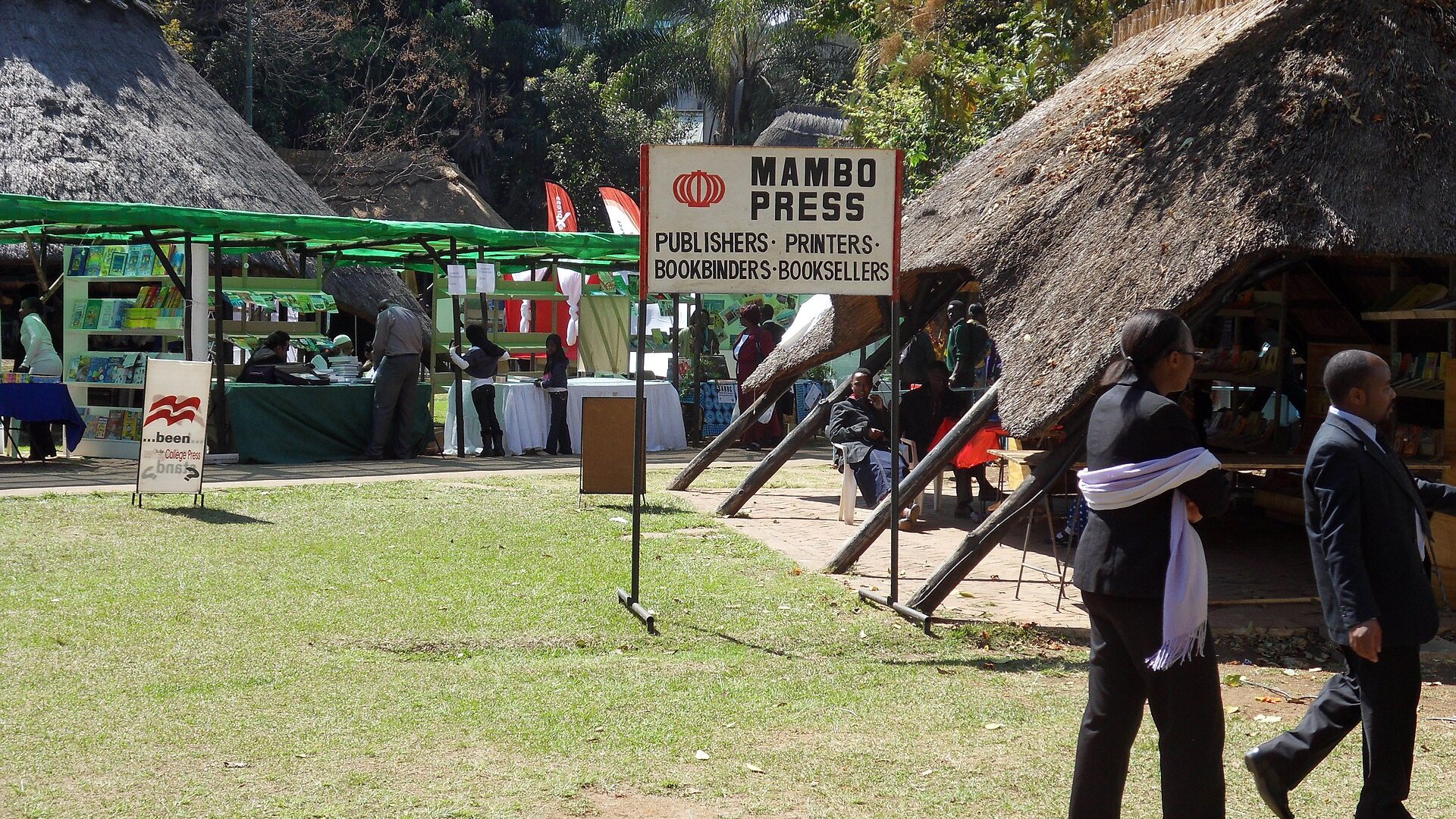 Thatched book stalls and a signboard beside a lawn at the Zimbabwe Book Fair, with visitors walking past