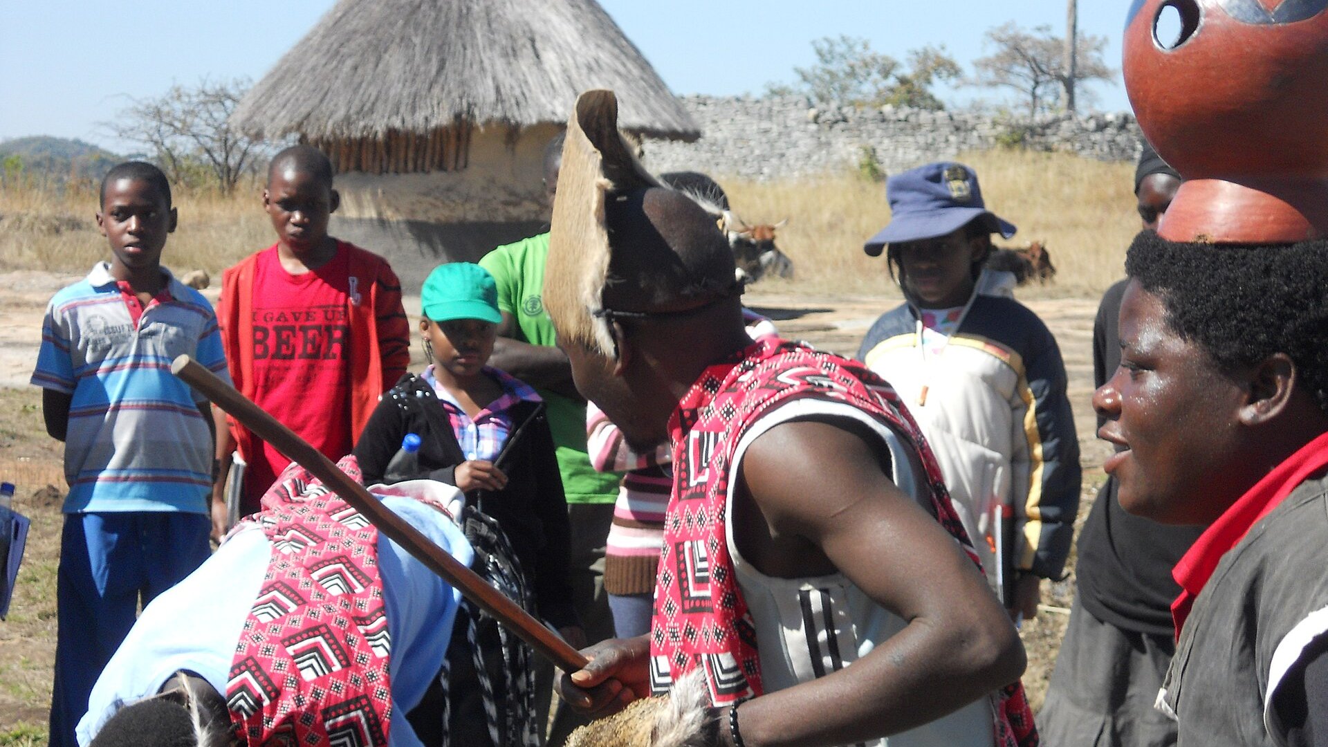 A dancer in an animal-skin headdress and red patterned cloth performs in front of onlookers beside a thatched hut and a dry-stone wall