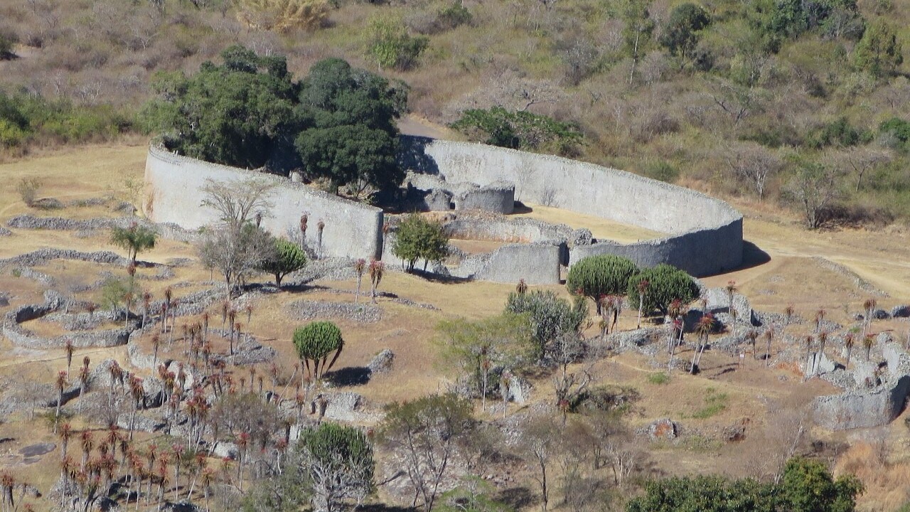 Aerial view of Great Zimbabwe with a long curved granite wall among trees, smaller stone enclosures and aloes on dry ground below a hillside