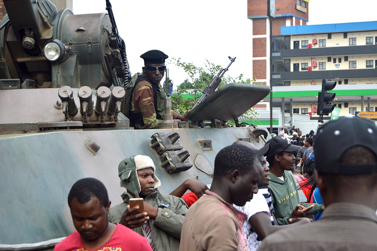 A soldier in a beret and sunglasses sits on an armoured vehicle in central Harare while civilians crowd around it