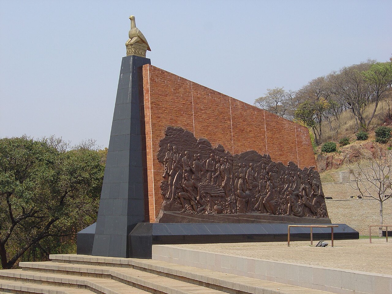 A bronze relief of liberation-war scenes on a brick wall beside a black obelisk topped by a golden Zimbabwe bird at Heroes' Acre, Harare